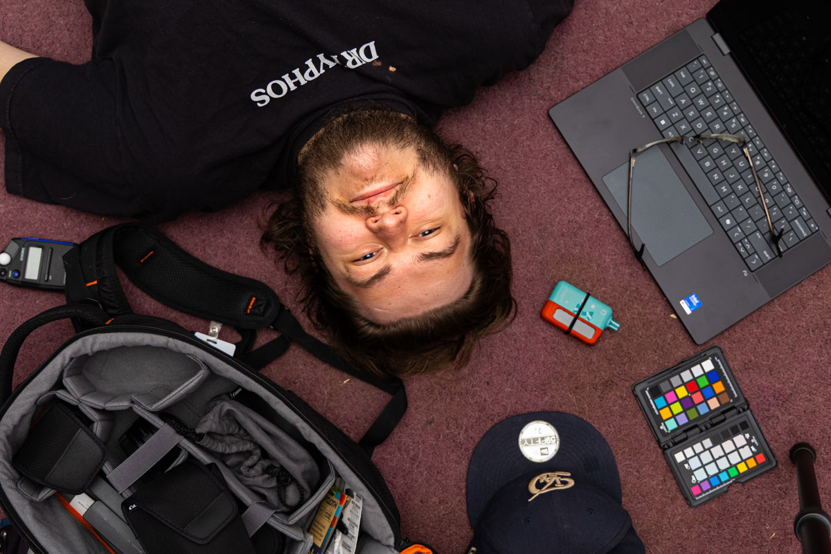 A man laying on the floor. He is looking at the camera placed above him looking down. He is surrounded by some photography gear, a laptop, and a few personal effects.