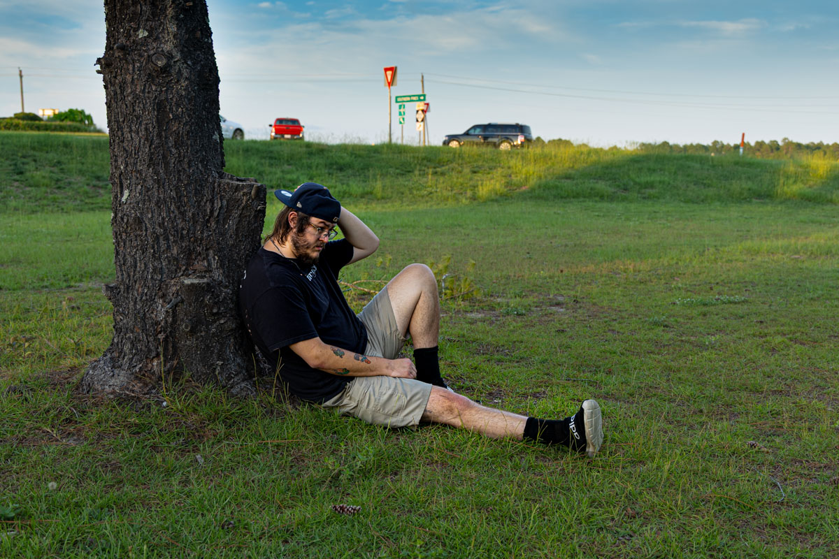 A man is sitting next to a tree in a field. His back is leaning on the tree and his left hand is fixing his hat. He looks midly dissapointed.