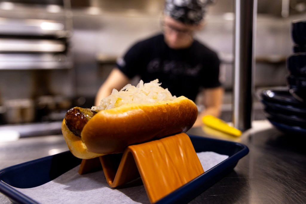 A bratwurst dog on a tray sitting in a service window of a commercial restaurant and the cook who made it in the background.
